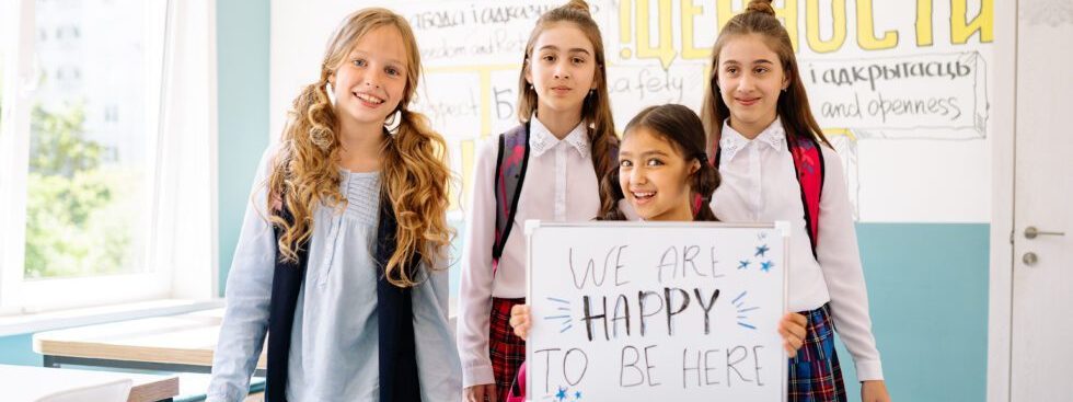 Four Female Students Holding A Whiteboard Stating 'We Are Happy To Be Here'